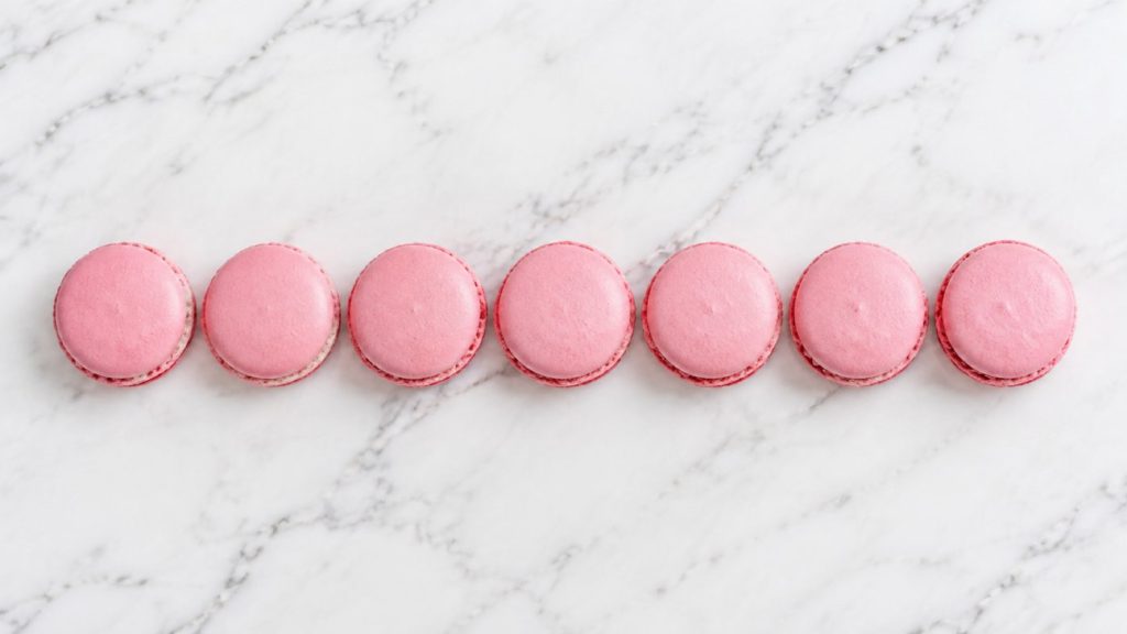 Seven pink macarons in a single row on white marble, photographed from above.